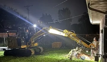 Excavator and crew working beside a brick home after dark under work lights