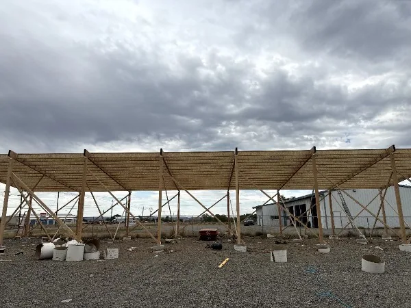 Long timber pole structure framed on a gravel pad under a cloudy sky